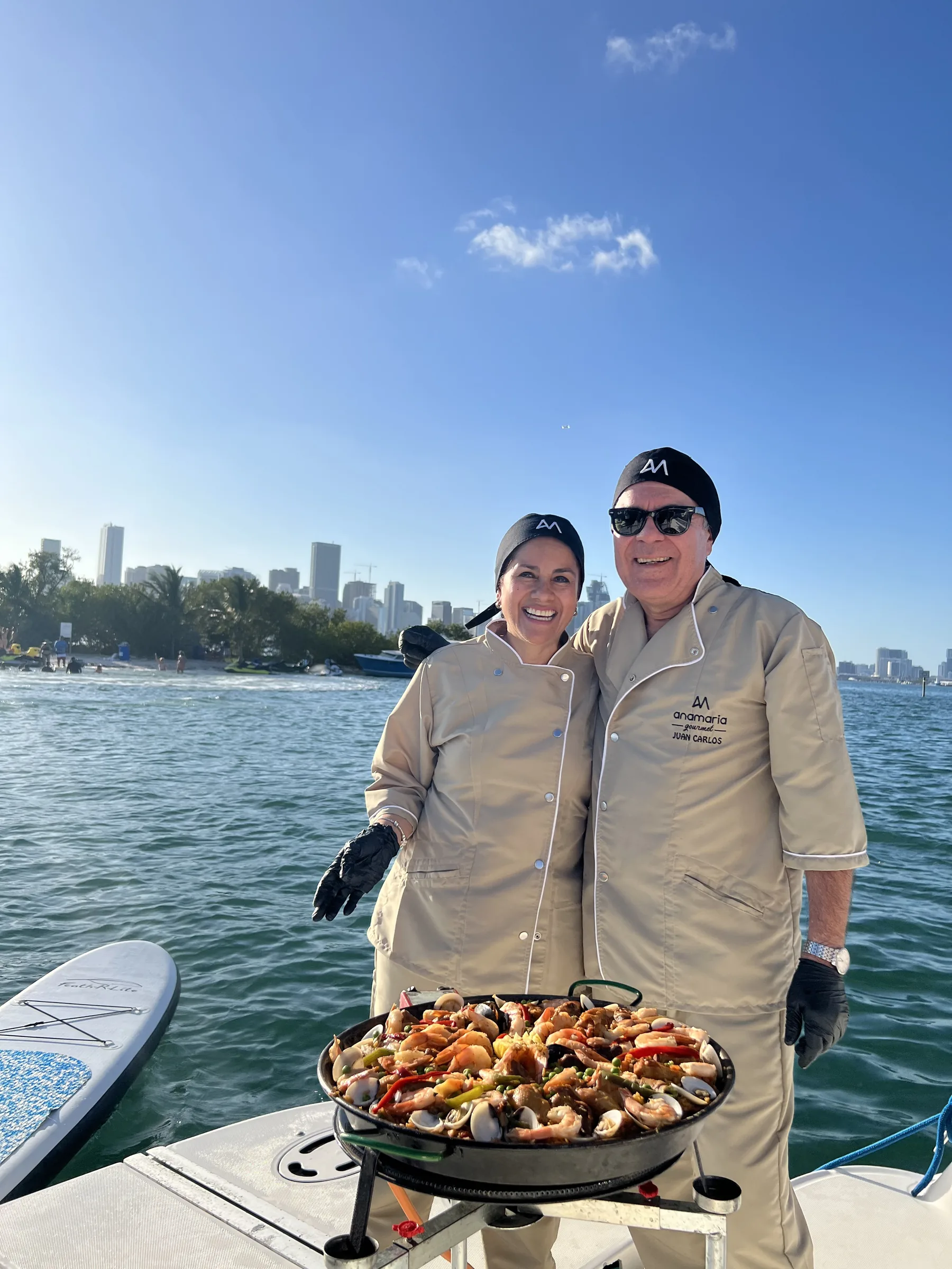 Ana y Juan con uniforme de chef sirven una paella marinera en un bote, con el horizonte de Miami al fondo